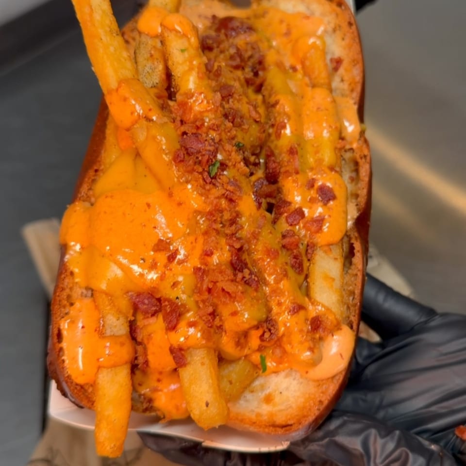 Gloved hands hold a loaded hot dog bun overflowing with french fries, cheese sauce, and bacon bits.
