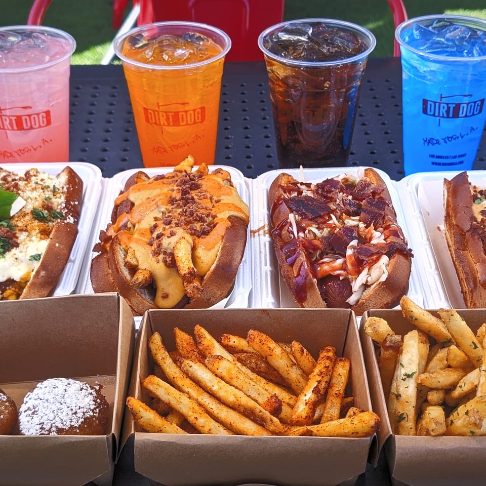 Four Dirt Dog hot dogs, three fries, and two donut holes with four colorful drinks on a black table.