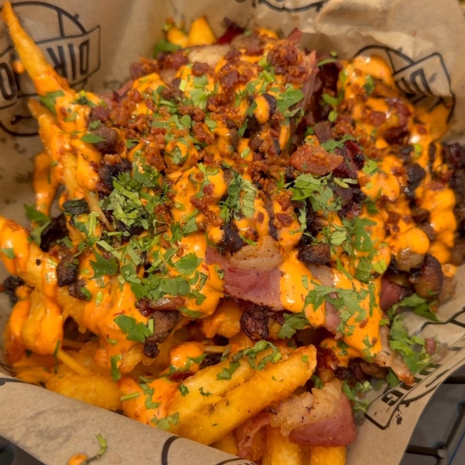 Loaded fries topped with orange sauce, crispy bacon, grilled meat, and chopped herbs in branded paper serving trays.