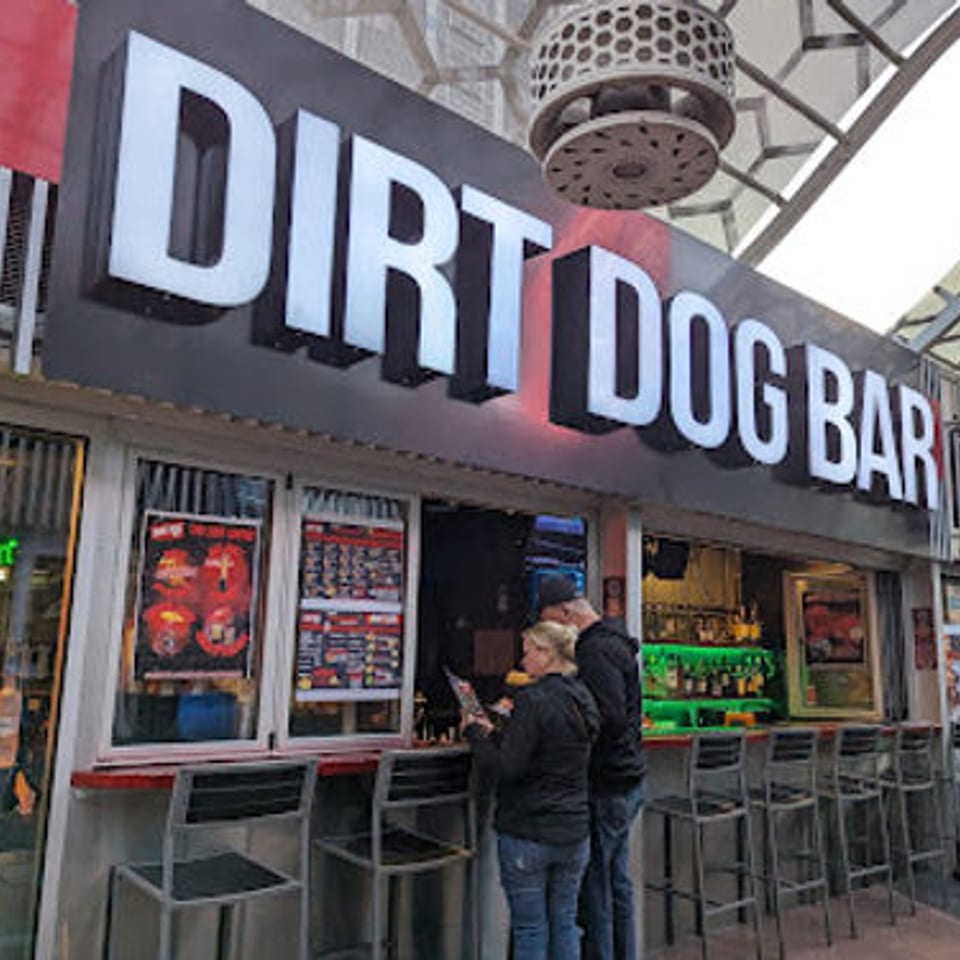 Exterior view of Dirt Dog Bar with a large sign, bar stools, and two people standing at the counter.