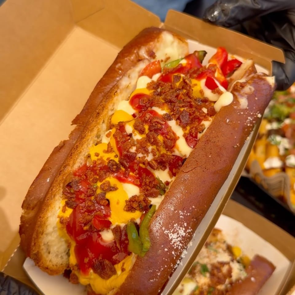 A gourmet hot dog in a pretzel bun, topped with cheese, bacon, peppers, and sauces.