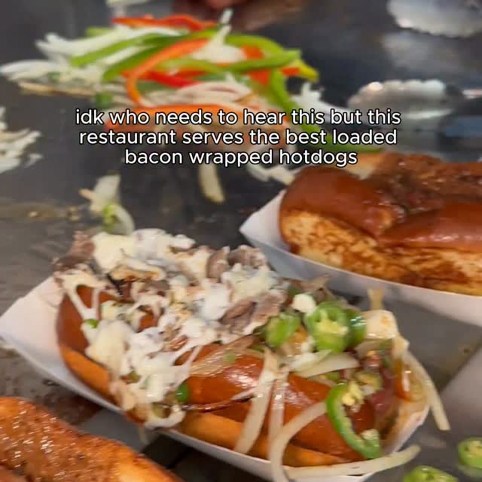 Loaded bacon-wrapped hotdogs topped with onions, peppers, sauces, and jalapeños sit in trays on a restaurant griddle.