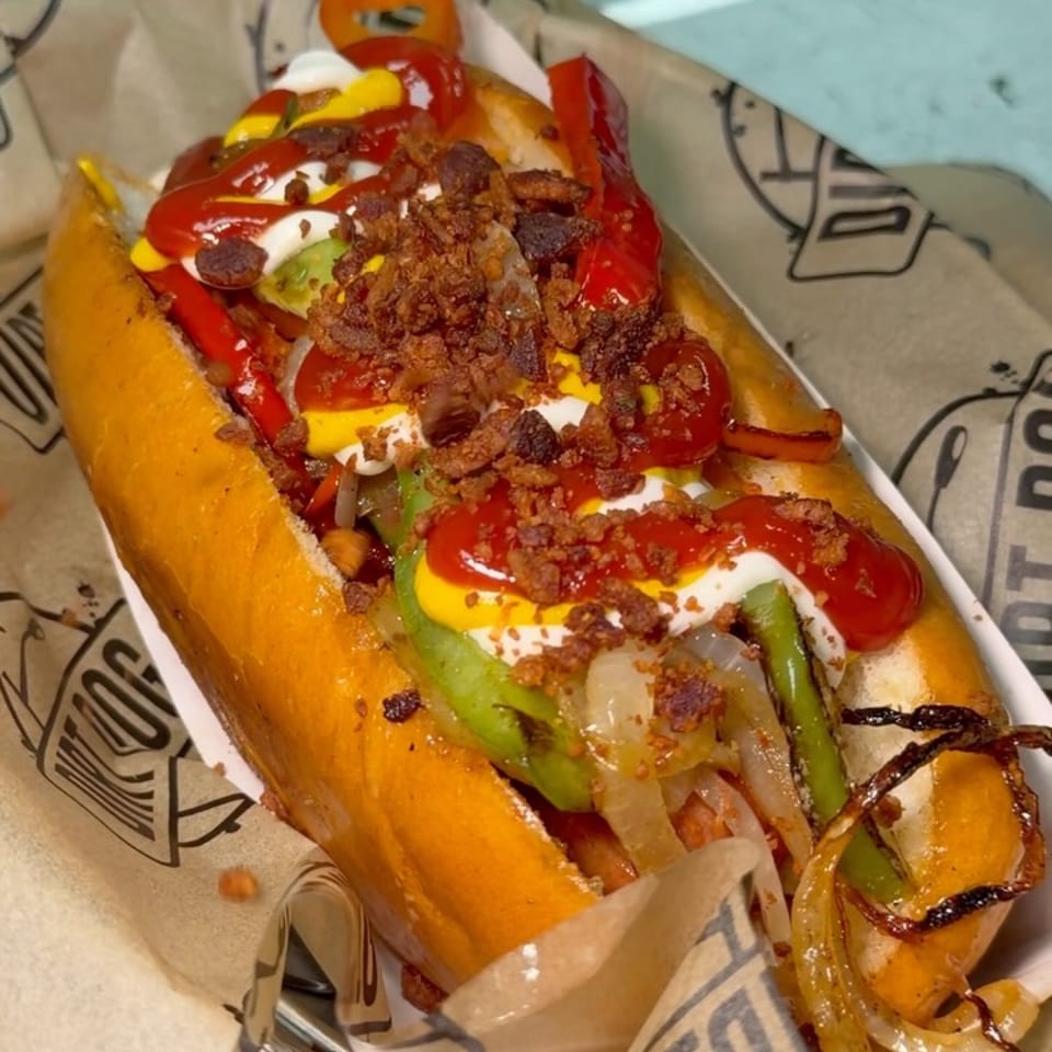 Loaded hot dog topped with ketchup, mustard, mayonnaise, crispy bacon, peppers, onions, and jalapeños on branded paper.