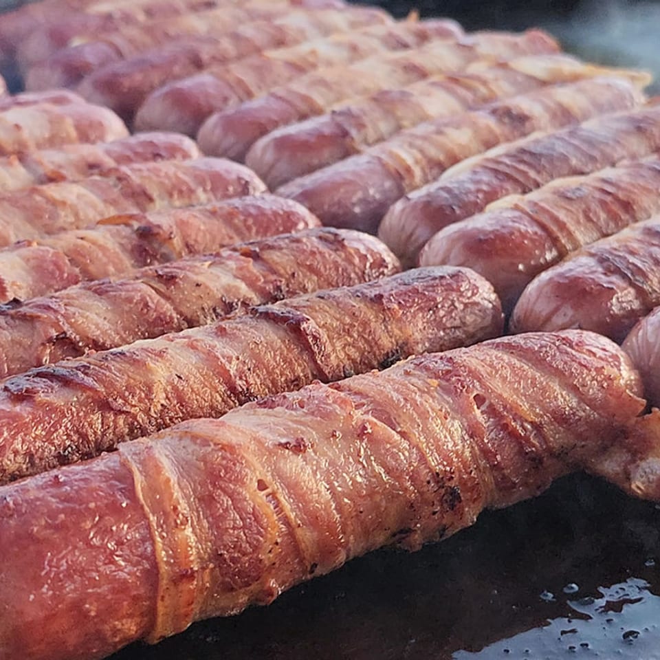 Rows of bacon-wrapped sausages grilling on a hot, oiled flat-top griddle.