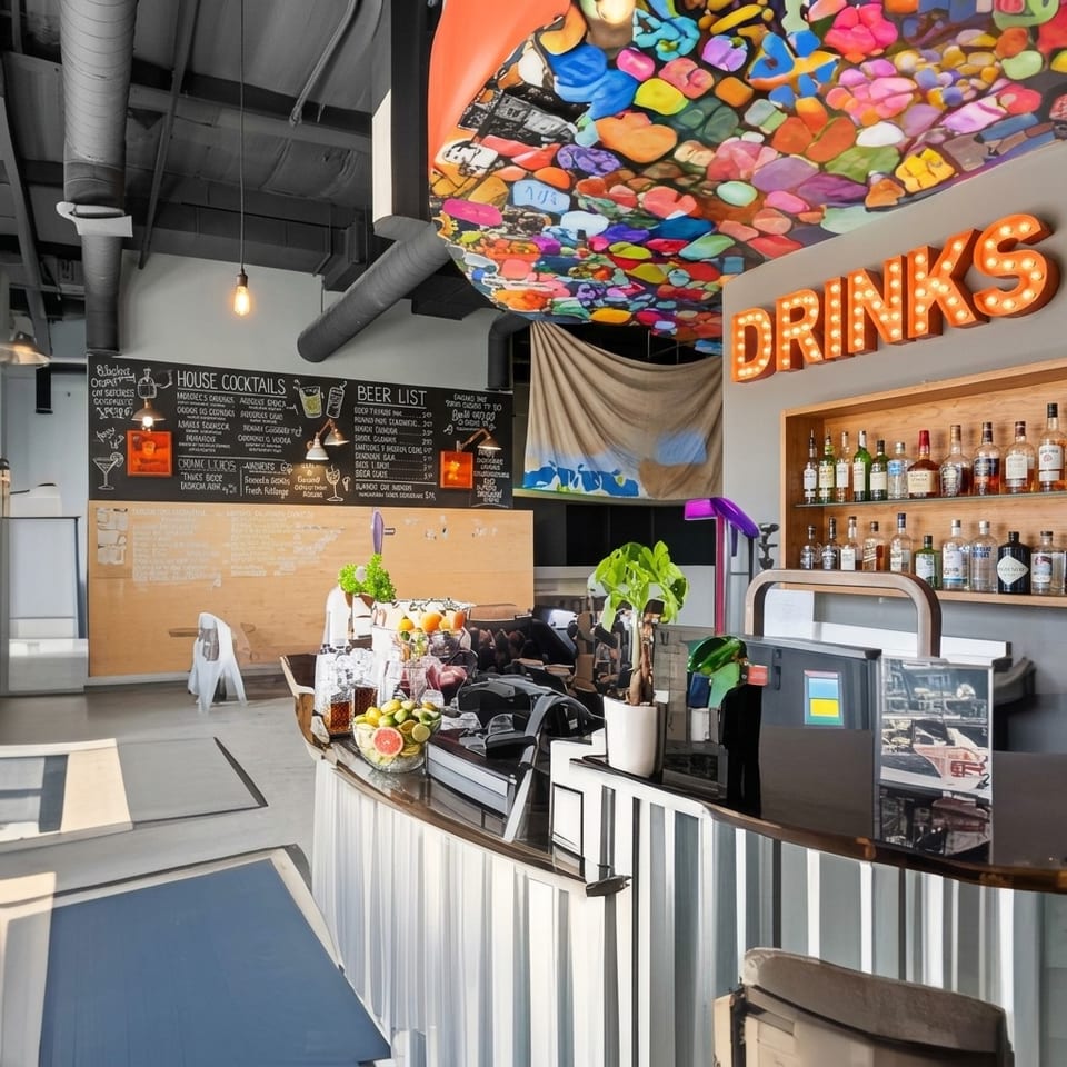Colorful cocktail bar with illuminated DRINKS sign, liquor shelves, chalkboard menus, exposed ducts, and vibrant ceiling artwork.