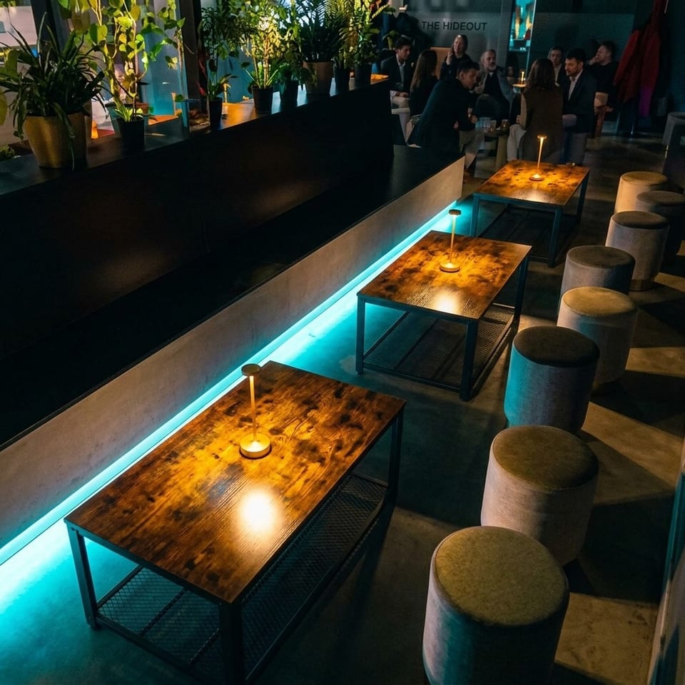 Dimly lit lounge with wooden tables, candle lamps, upholstered stools, potted plants, and guests socializing beneath a Hideout sign.
