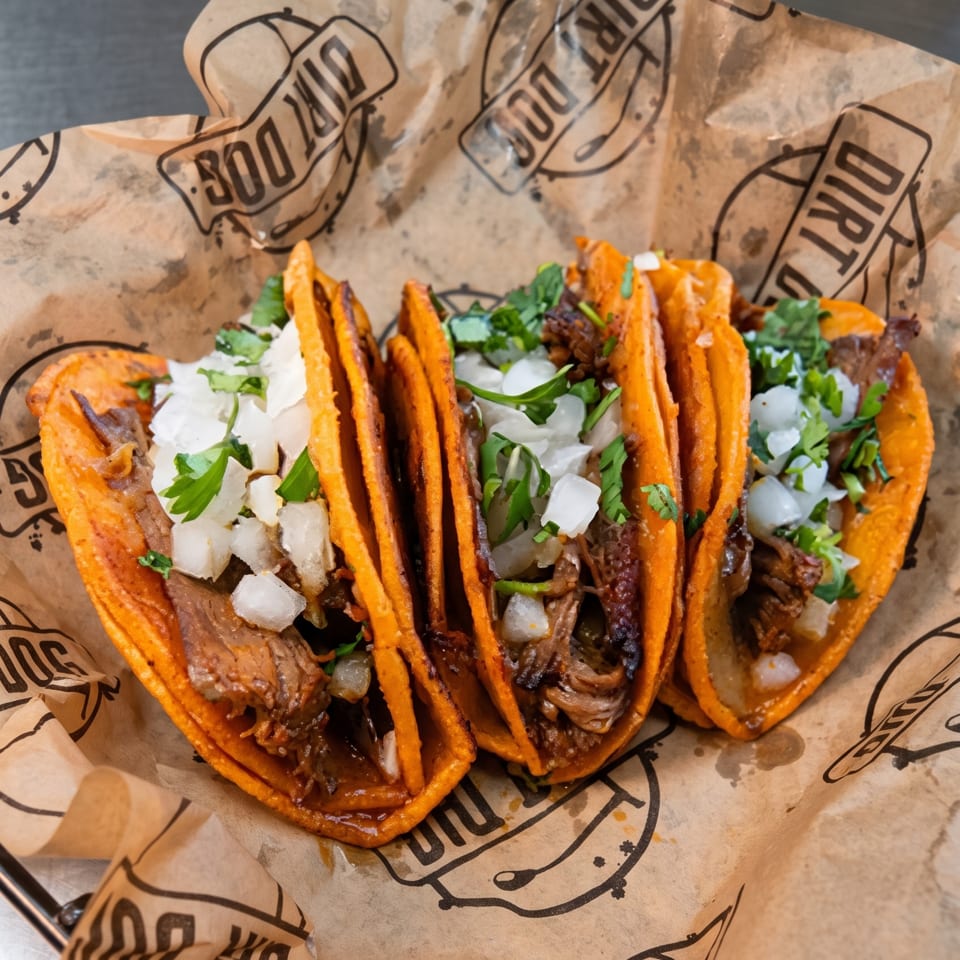 Three orange tacos filled with grilled meat, diced onions, cilantro, and creamy sauce, served on branded paper.