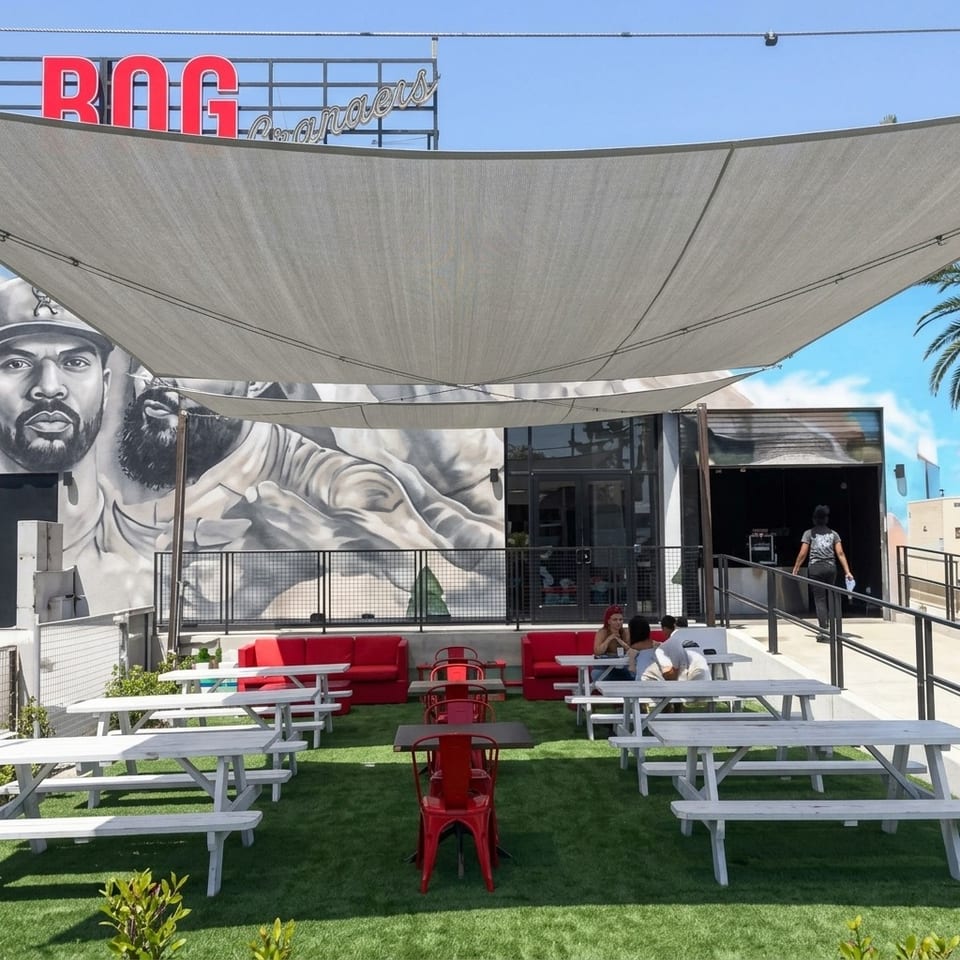 Outdoor ROG Promoters restaurant patio with picnic tables, red seating, shade sails, and a large black-and-white mural.