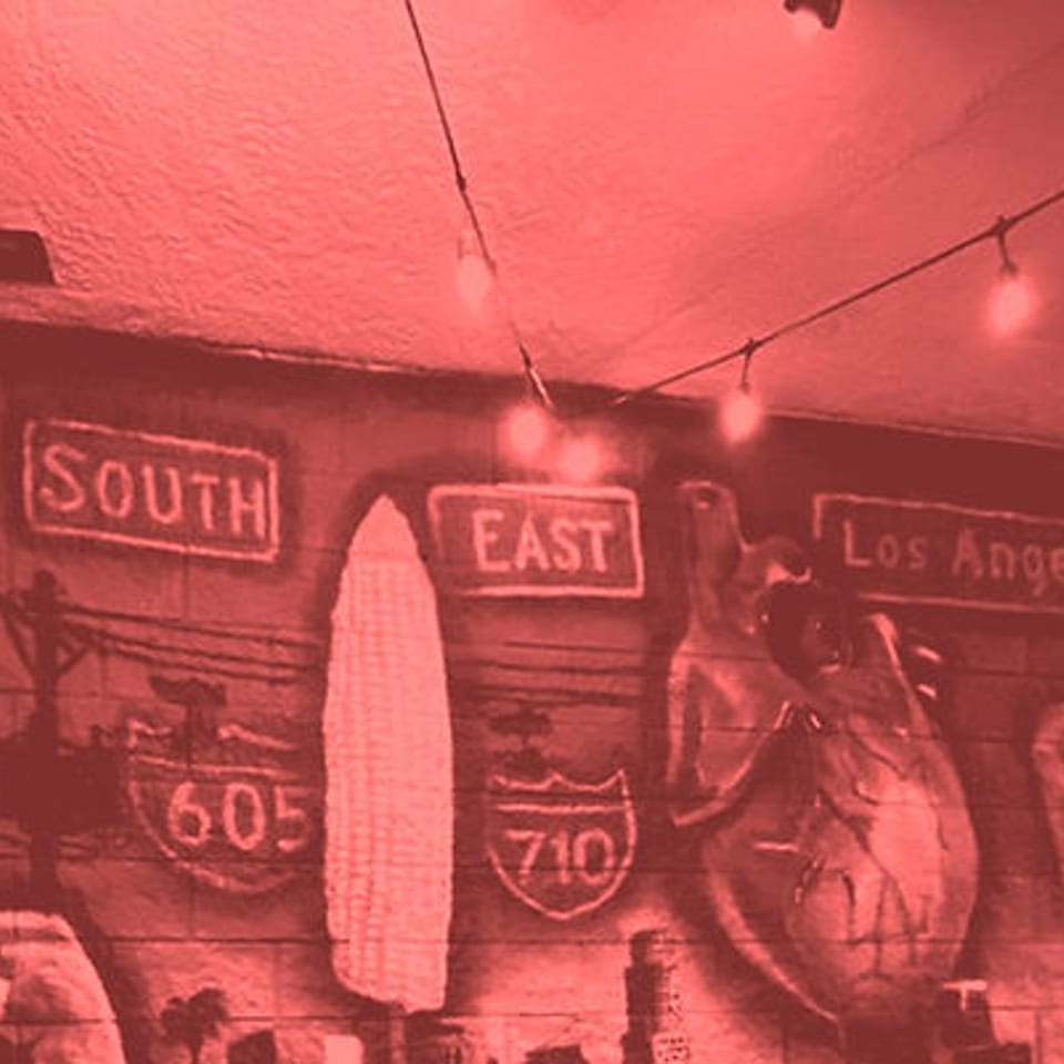 A red-tinted mural with "South," "East," "Los Angeles," and freeway signs, under string lights.