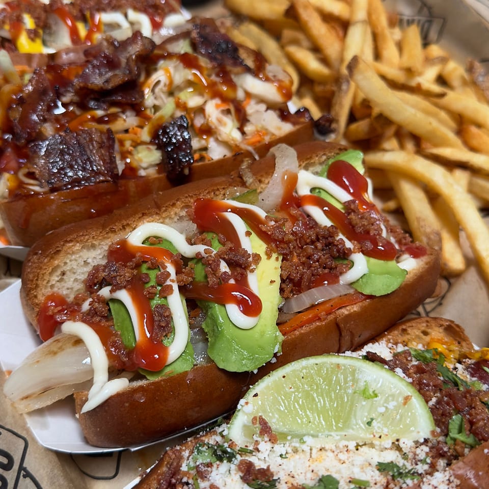 A platter of gourmet hot dogs with various toppings, including avocado, bacon, and sauces, alongside french fries.